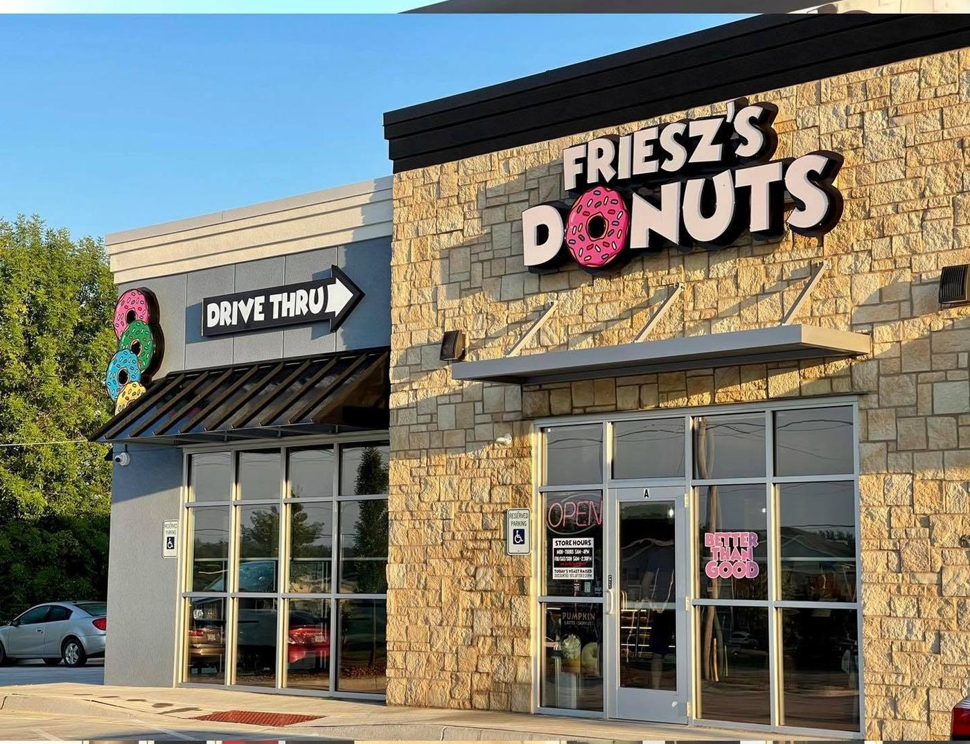The shop from the parking lot: a stone-faced building with Friesz's Donuts in dimensional letters, a pink sprinkled donut standing in for the O, a drive thru sign with three stacked donuts, and a neon open sign in the window.